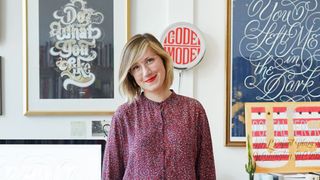 Jessica Hische headshot, woman in a red top in front of her lettering work